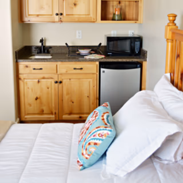 A cozy room featuring a bed with white bedding and a colorful decorative pillow, adjacent to a small kitchenette area with wooden cabinets, a black faucet and sink, a microwave, and a mini refrigerator.