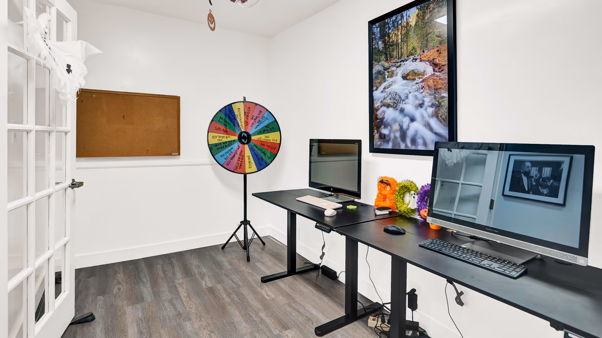 A small office or activity room with two computer monitors on a long black desk, a colorful prize wheel on a stand, a cork bulletin board on the wall, and a framed nature photograph above the desk. The room has white walls, wood flooring, and a glass-paneled door.