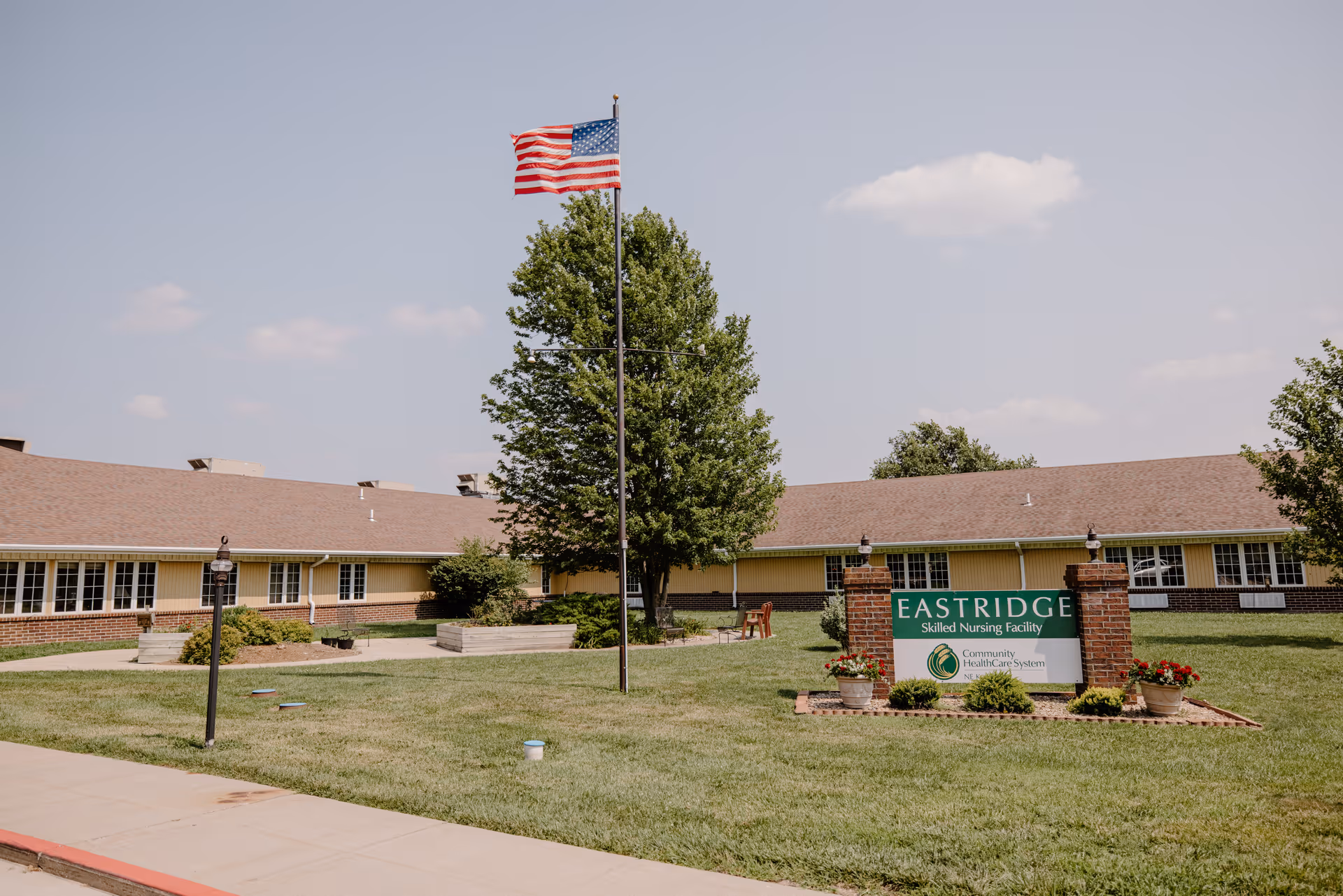 Exterior view of Eastridge Skilled Nursing Facility with a large green lawn, an American flag on a flagpole, and a sign displaying the facility's name and affiliation with Community HealthCare System. The building is single-story with a brown roof and beige walls, surrounded by trees and shrubs under a partly cloudy sky.
