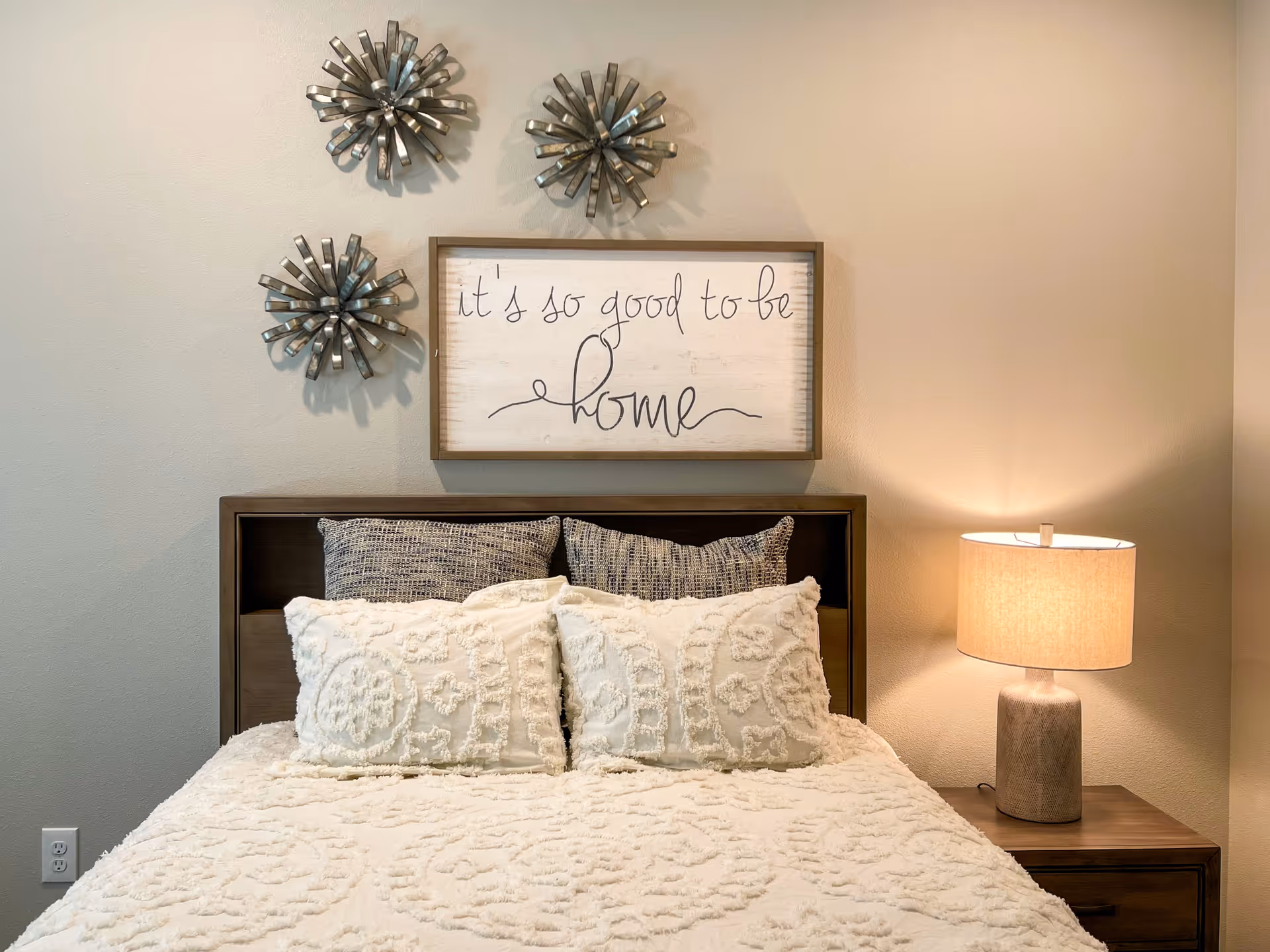 A cozy bedroom with a bed featuring textured white bedding and pillows. Above the headboard, there is a framed sign that reads 'it's so good to be home' and three decorative metal wall sculptures. A wooden nightstand with a beige lamp is positioned to the right of the bed.