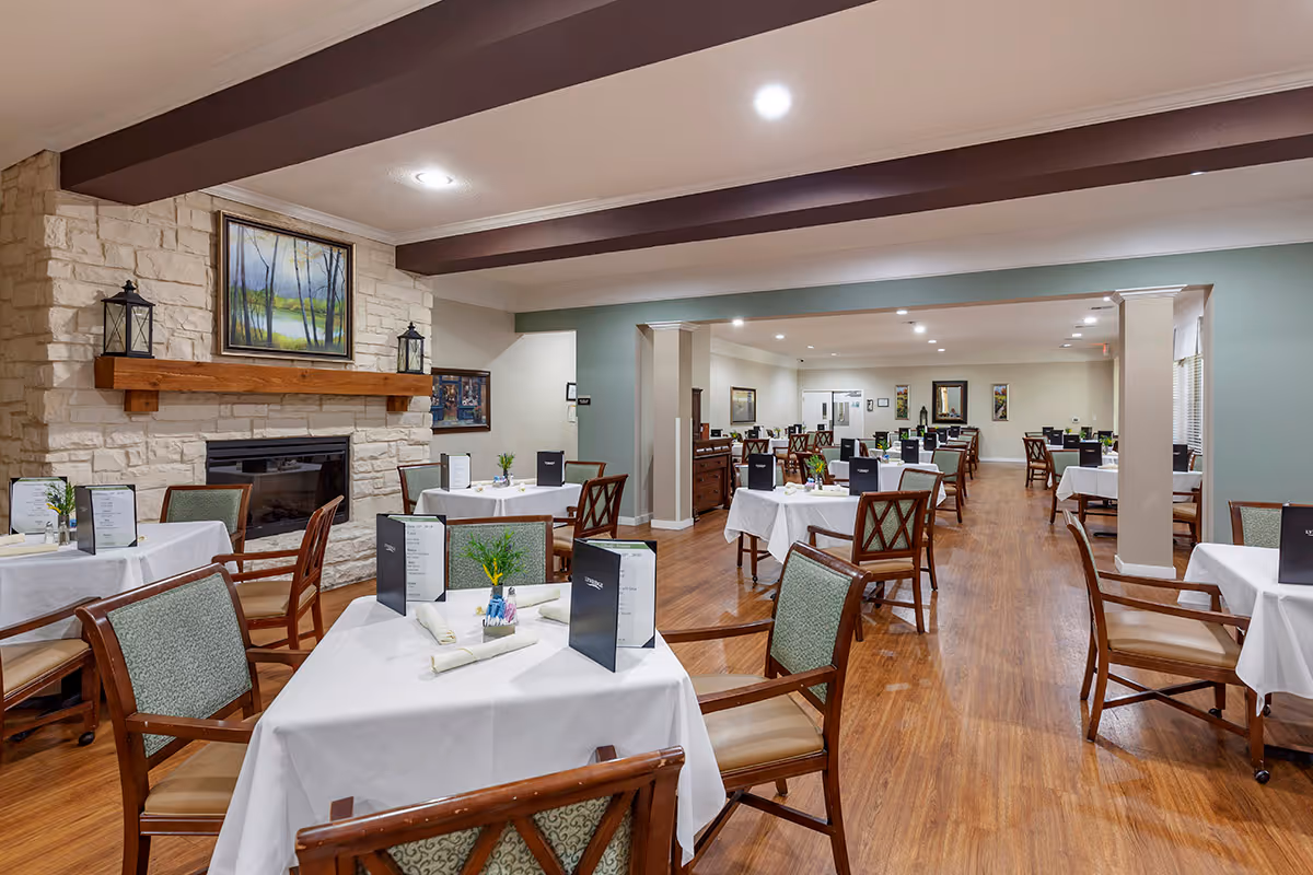 A spacious dining room with multiple tables covered in white tablecloths, each set with menus, napkins, and small flower arrangements. The room features wooden floors, a stone fireplace with a wooden mantle, and a painting above the fireplace. The ceiling has exposed wooden beams and recessed lighting.