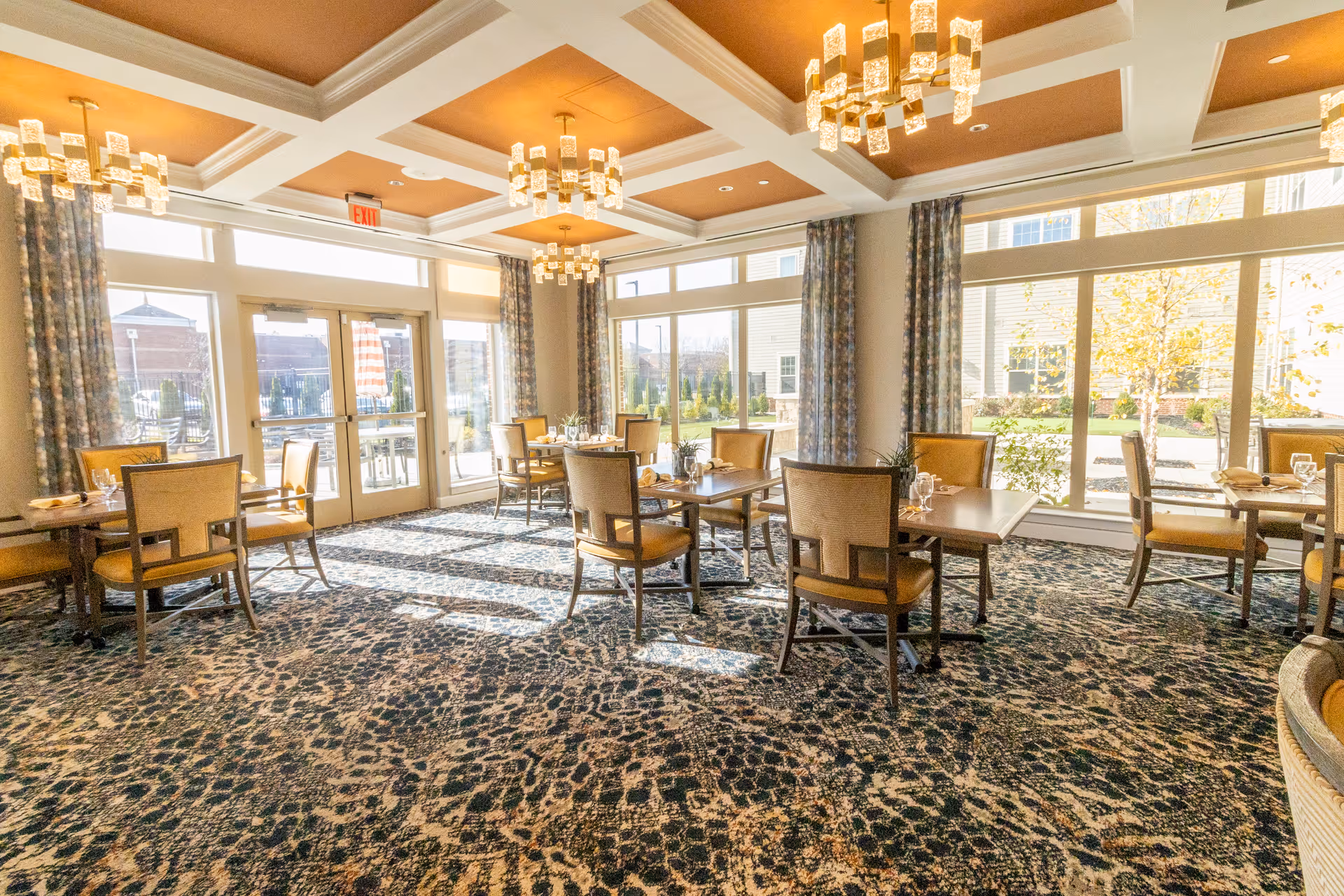 A bright dining room with several tables and chairs arranged neatly. The room features large windows with floral curtains allowing natural light to fill the space. The ceiling has recessed panels with modern chandeliers hanging down. The carpet has a patterned design, and the tables are set with glasses and napkins.