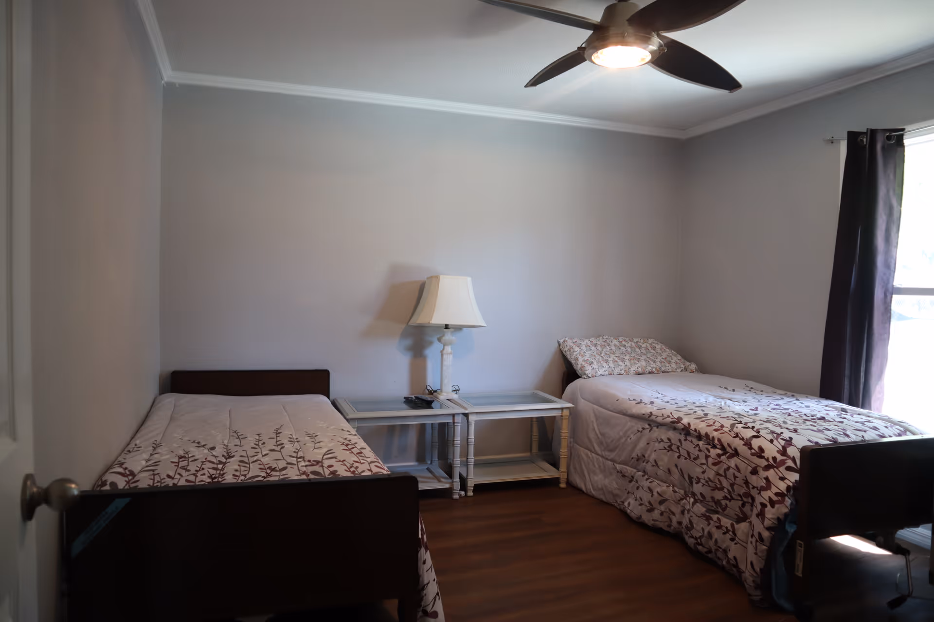 A simple bedroom with two single beds placed against opposite walls. Each bed has a floral-patterned comforter. Between the beds is a white nightstand with a lamp on it. The room has light gray walls, a ceiling fan with a light, and a window with dark curtains letting in natural light.