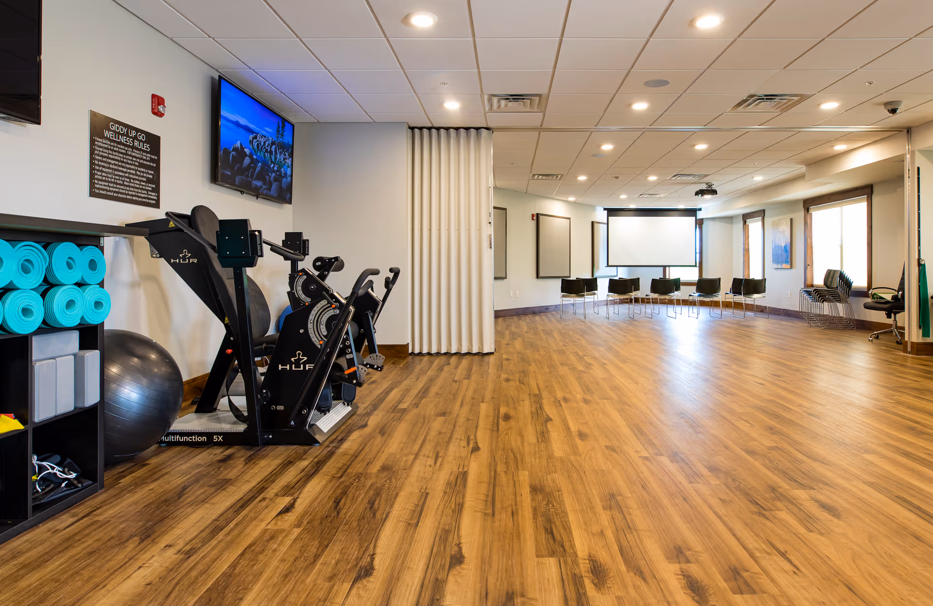 A spacious wellness room with wooden flooring featuring exercise equipment including stationary bikes, yoga mats, and an exercise ball on the left side. A television is mounted on the wall above the equipment, and a sign with wellness rules is visible. The room has a large open area with chairs arranged in a semi-circle near a projector screen at the far end. Large windows on the right side allow natural light to enter.