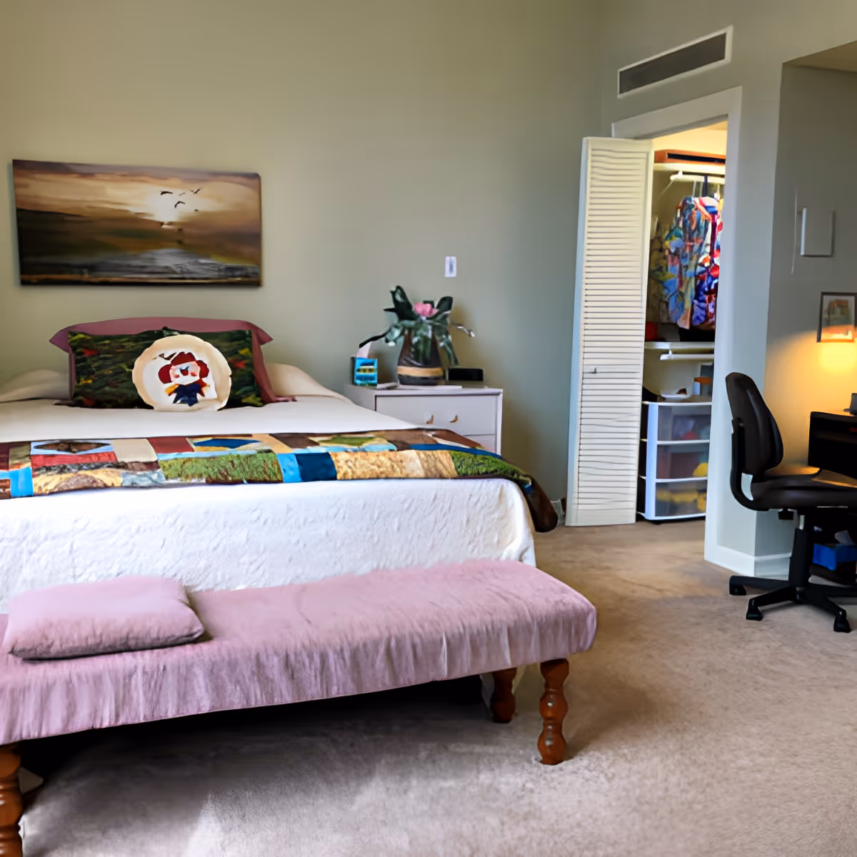 A cozy bedroom with a bed covered in a white quilt and a colorful patchwork blanket. There is a pink cushioned bench at the foot of the bed with a matching pillow. On the bed are two decorative pillows, one with a scenic design and another with a circular pattern. A nightstand next to the bed holds a potted plant and some small items. The room has light green walls, a painting of birds flying over a landscape above the bed, and an open closet door revealing hanging clothes. A black office chair is positioned near a desk with a lamp on the right side of the room.