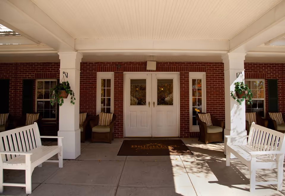 Covered brick front entrance with white double doors, white benches, hanging plants and seating on a porch.
