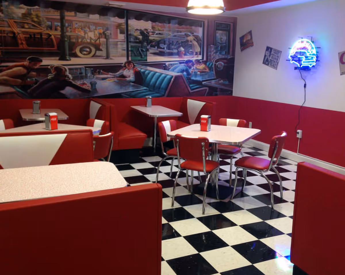 Retro-style dining room with red booths and chairs, black-and-white checkered floor, wall mural and neon sign.