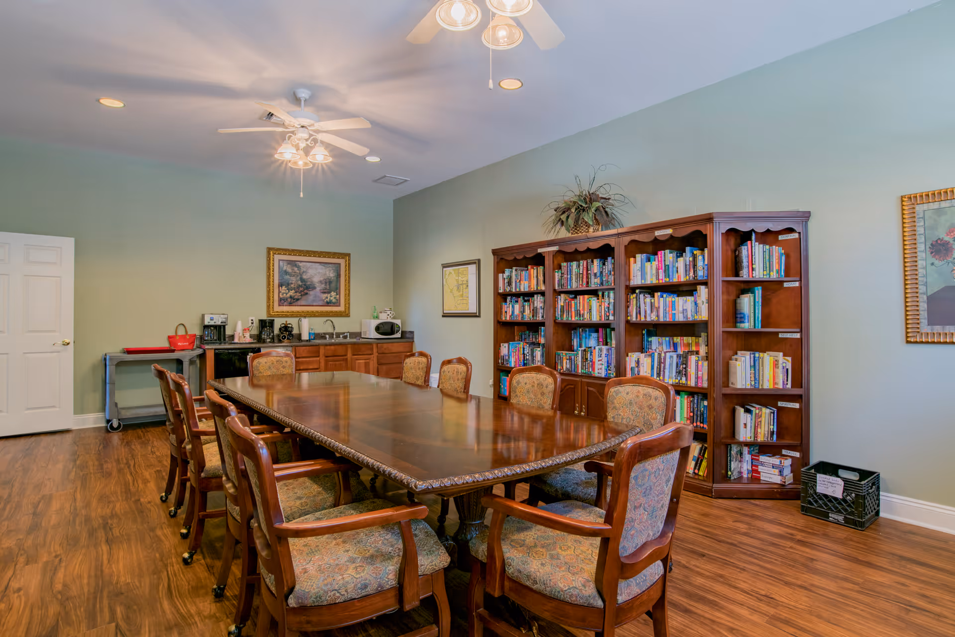 A well-lit room with a large wooden table surrounded by eight cushioned chairs. Against the wall is a wooden bookshelf filled with books and a small plant on top. The room has light green walls, wooden flooring, a ceiling fan with lights, and framed artwork on the walls. There is also a small kitchenette area with a microwave, coffee maker, and sink.