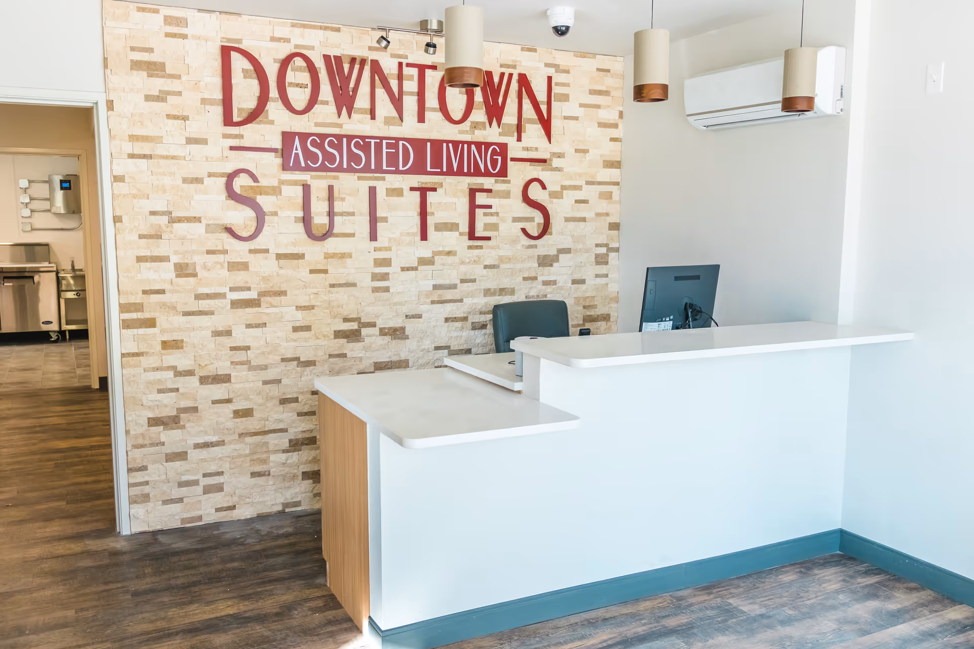 Reception area of Downtown Assisted Living Suites with a white reception desk, a computer monitor, and a stone accent wall featuring the facility's name in large red letters. The floor has wood-style flooring and there are pendant lights hanging from the ceiling.