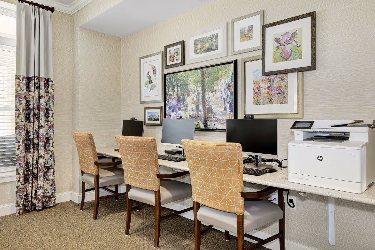 A small computer workstation area with three chairs and three desktop computers on a long counter. There is a white HP printer on the right side of the counter. The wall behind the computers is decorated with multiple framed paintings and artwork. A window with floral-patterned curtains is on the left side of the room.