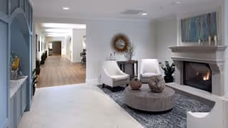 Well-lit senior living common area with a fireplace, upholstered chairs, round coffee table, and a hallway leading to other rooms.