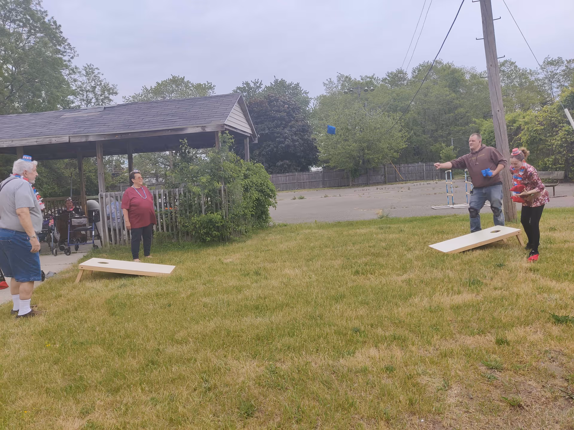A group of people playing cornhole on a grassy area near a wooden gazebo. Two cornhole boards are set up on the grass, and one person is in the act of tossing a bean bag. Trees and a fenced area are visible in the background under an overcast sky.