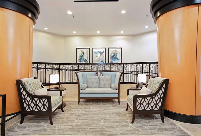 A seating area in a senior living facility featuring a light blue loveseat with two matching cylindrical pillows, flanked by two armchairs with patterned cushions. The chairs and loveseat have dark wooden frames with a circular lattice design on the sides. Two small round tables with lamps are positioned beside the armchairs. The area is carpeted with a light patterned rug and is framed by two large orange columns. Three abstract paintings hang on the wall behind a decorative black railing.