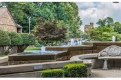 Outdoor area with multiple tiered stone fountains surrounded by greenery, bushes, and trees. There is a stone bench in the foreground and a brick building partially visible on the left side under a partly cloudy sky.