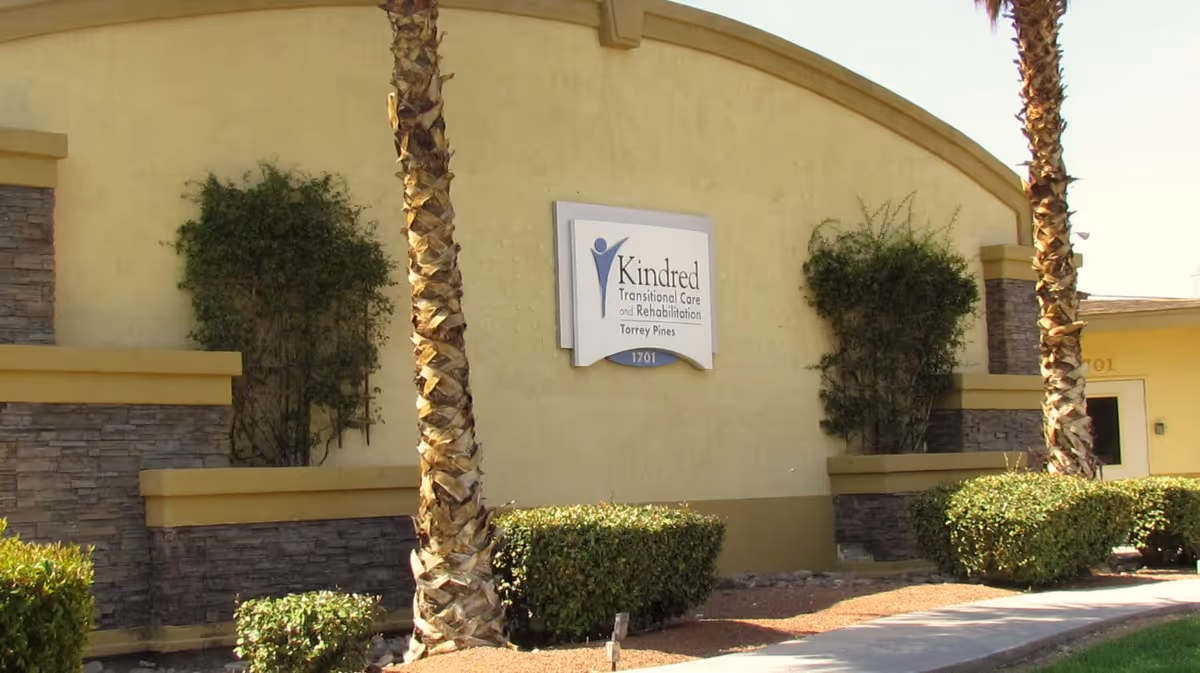 Exterior view of a building with a sign that reads 'Kindred Transitional Care and Rehabilitation Torrey Pines 1701'. The building has a beige and stone facade with two palm trees and trimmed bushes in front.