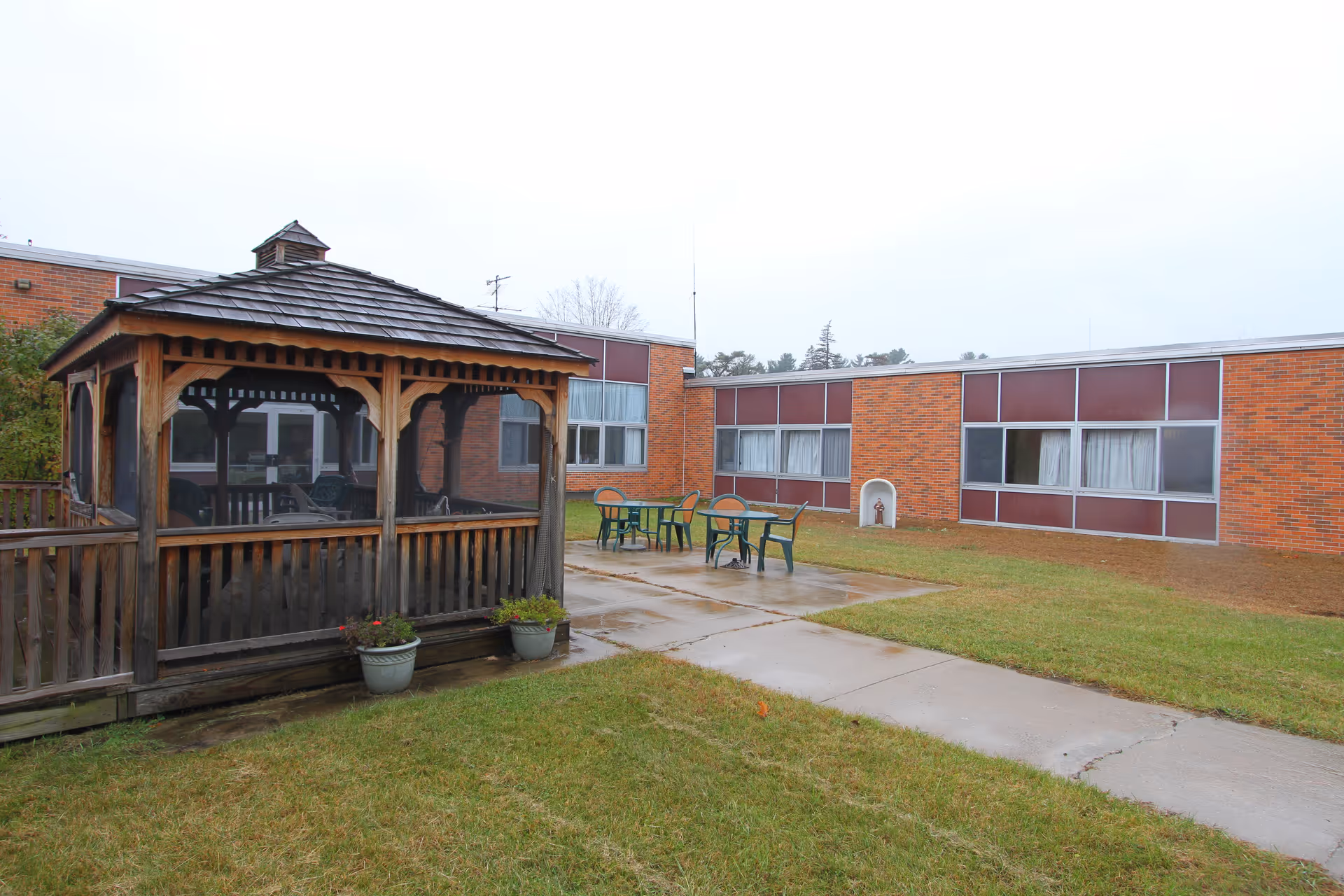 Outdoor courtyard with a wooden gazebo, patio tables, and a one-story brick building in the background.