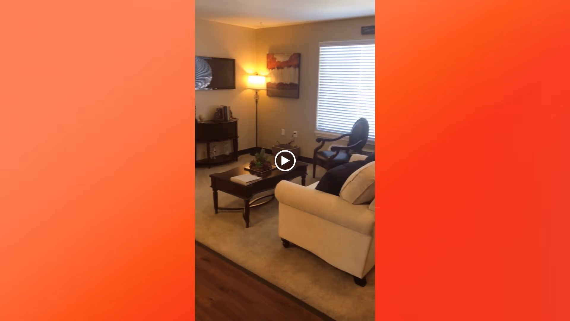 A cozy living room with a beige sofa, a wooden coffee table with decorative items, a wooden chair, a floor lamp, a wall-mounted TV, and a window with blinds letting in natural light.