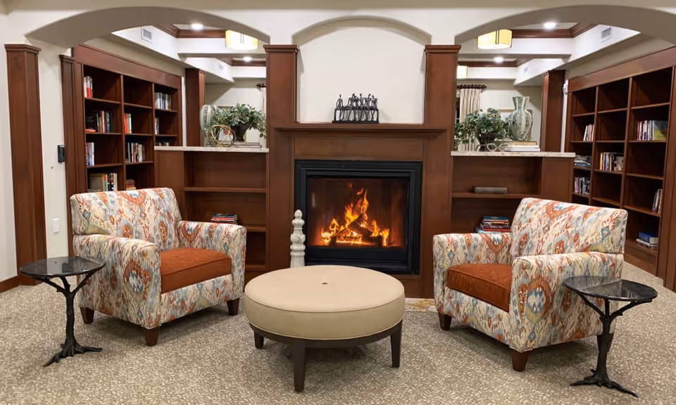 Cozy lounge with two patterned armchairs facing a lit fireplace, a round ottoman, side tables, and built-in bookshelves.