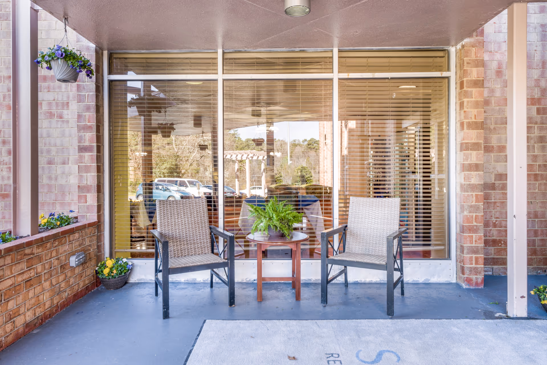 Outdoor seating area with two wicker chairs and a small round table with a potted fern, situated on a covered porch in front of large windows with blinds. The porch has brick walls and hanging flower pots with purple and yellow flowers.