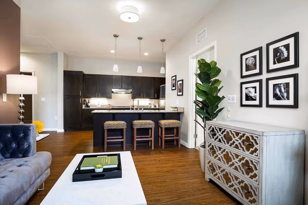 Open living room and kitchen with a sofa, marble coffee table, island with three stools, and decorative dresser with framed artwork.