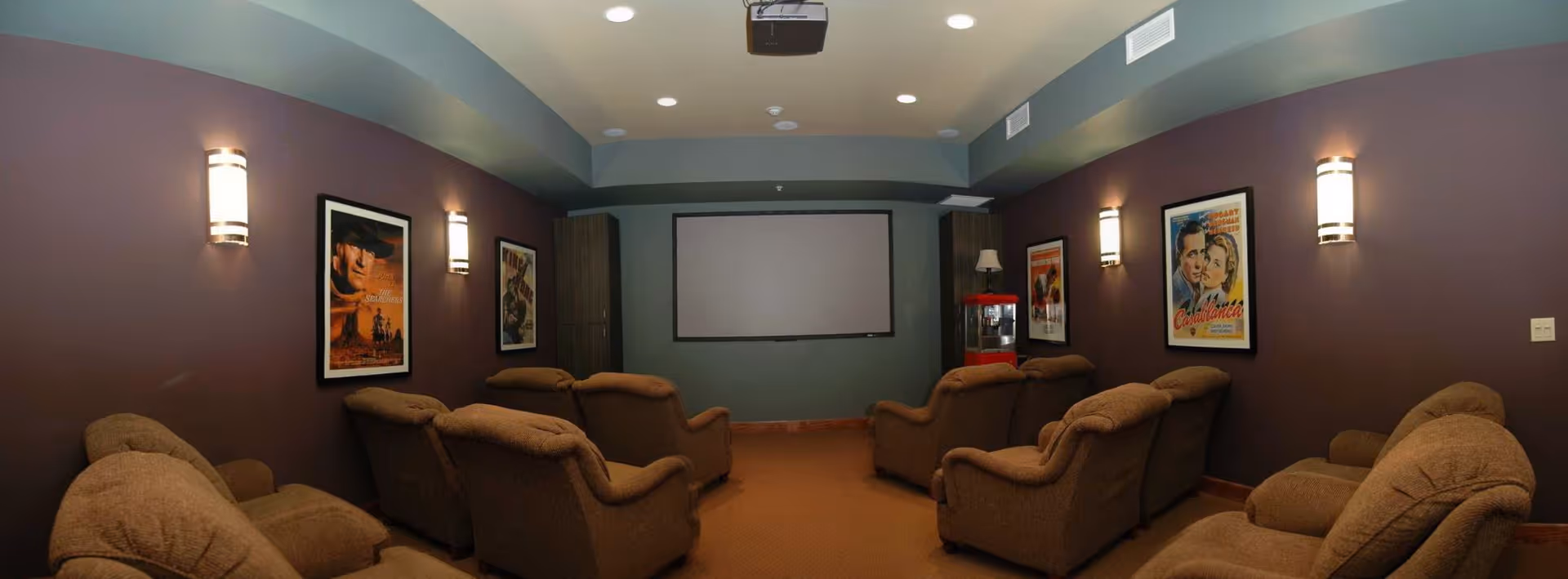 A cozy home theater room with brown upholstered reclining chairs arranged in rows facing a large projection screen. The walls are painted in a muted purple color and decorated with framed classic movie posters. Wall sconces provide soft lighting, and a popcorn machine is visible in the back corner of the room.
