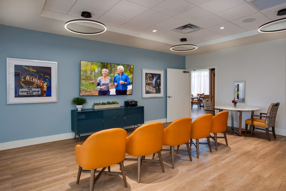 A modern lounge area with five orange chairs arranged in a row facing a wall-mounted flat screen TV displaying an elderly couple jogging. The wall behind the TV is painted blue and has two framed pictures on either side. Below the TV is a dark green cabinet with plants and electronic devices on top. To the right, there is a small round table with two plaid upholstered chairs and a vase with flowers. The room has wood flooring, circular ceiling lights, and an open door leading to another room with more chairs and tables.