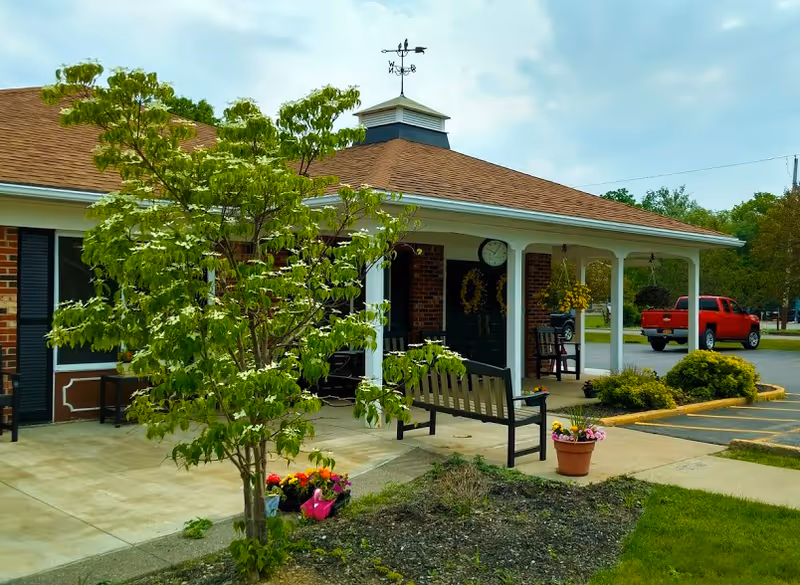 Front entrance of a brick senior living building with a covered porch, benches, potted flowers, and a small tree in the foreground.