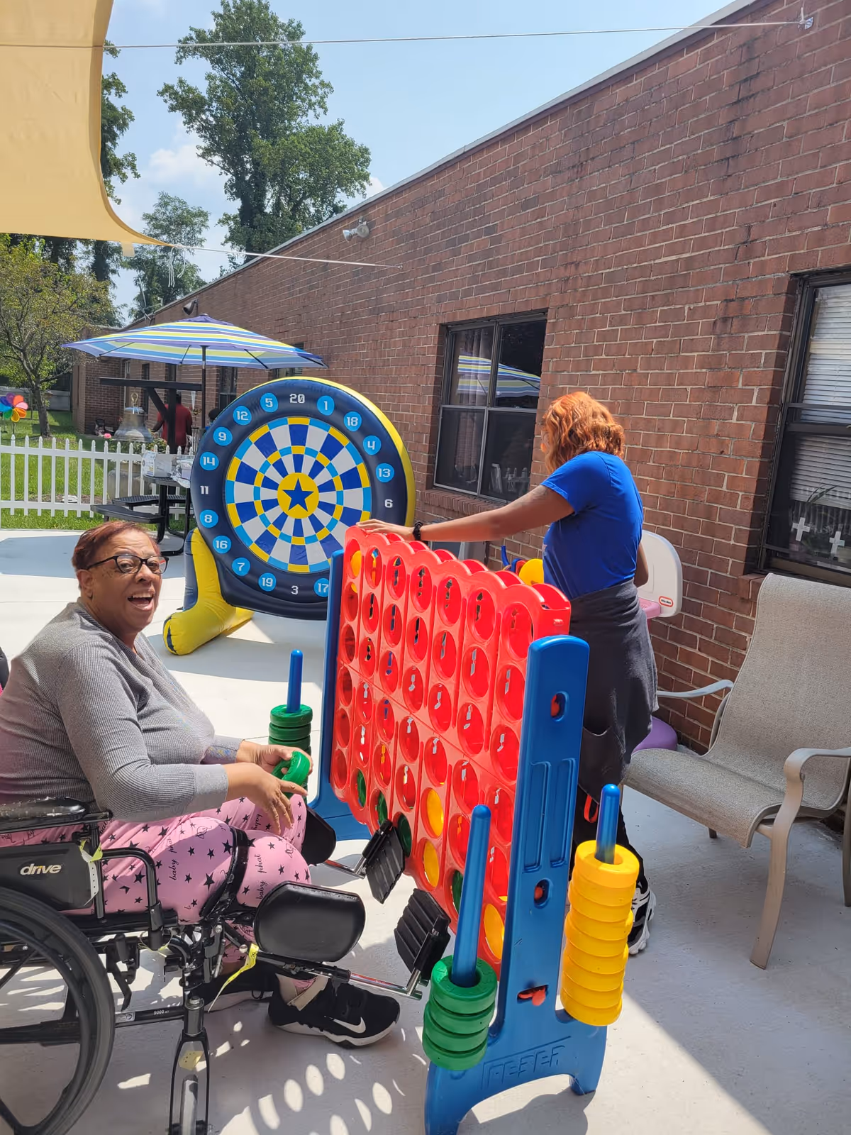 Two women playing a large outdoor Connect Four game on a patio. One woman is sitting in a wheelchair smiling, wearing glasses, a gray top, and pink pants with star patterns. The other woman, standing, is wearing a blue shirt and has reddish hair. Behind them is a large inflatable dartboard and a brick building wall with windows. There are outdoor chairs and a table with an umbrella in the background.