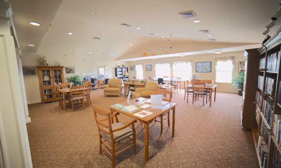 A spacious, well-lit common area in a senior living facility with multiple wooden tables and chairs arranged throughout the room. There are two beige armchairs near the center, bookshelves filled with books along the walls, and large windows letting in natural light. The carpet has a patterned design, and the ceiling has recessed lighting.
