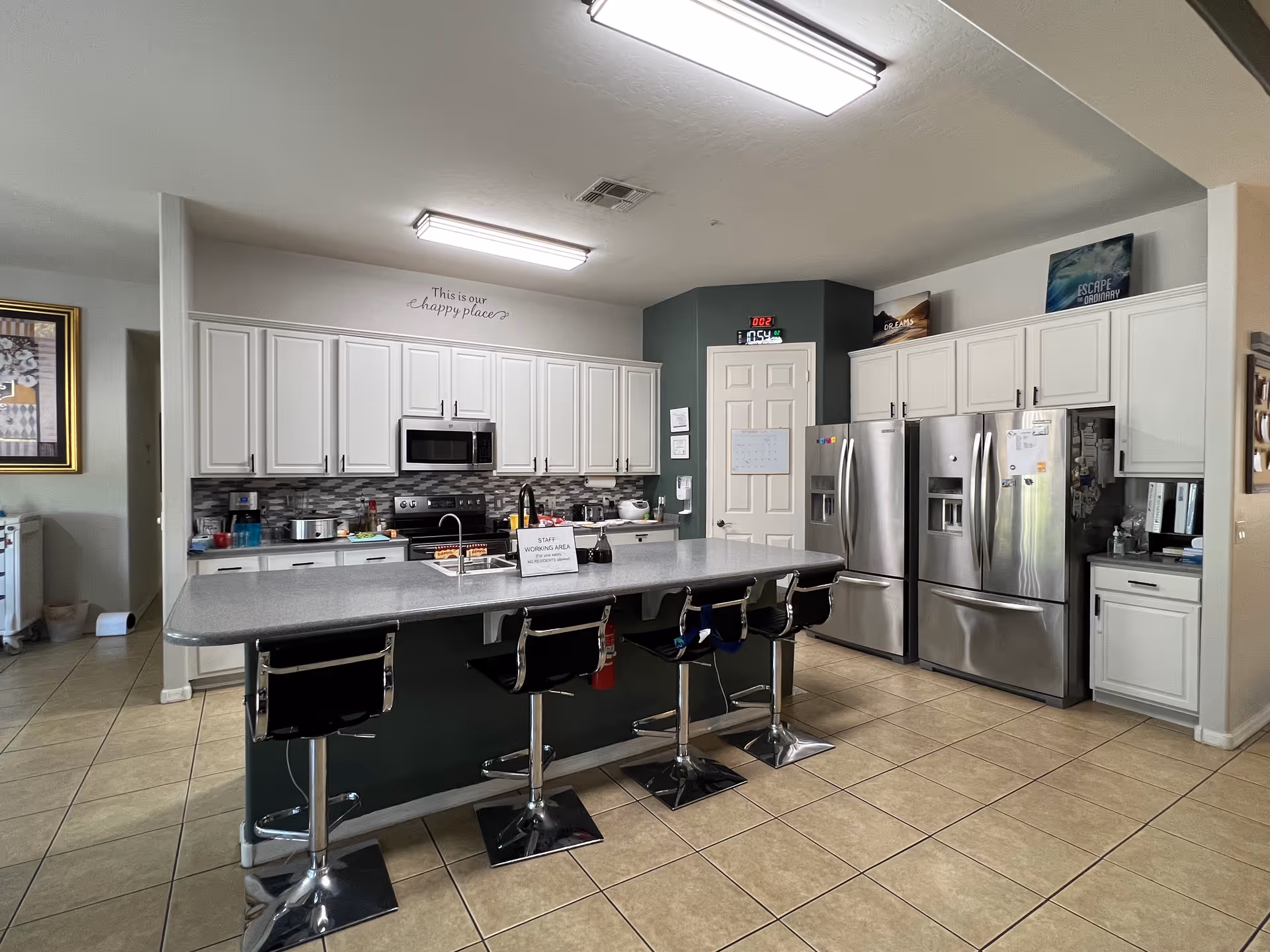 A modern kitchen with white cabinets, a large island with four black bar stools, two stainless steel refrigerators, a microwave, and a stove. The kitchen has tiled floors and bright overhead lighting. On the wall above the cabinets, there is a sign that reads 'This is our happy place'. A sign on the island indicates it is a staff working area.