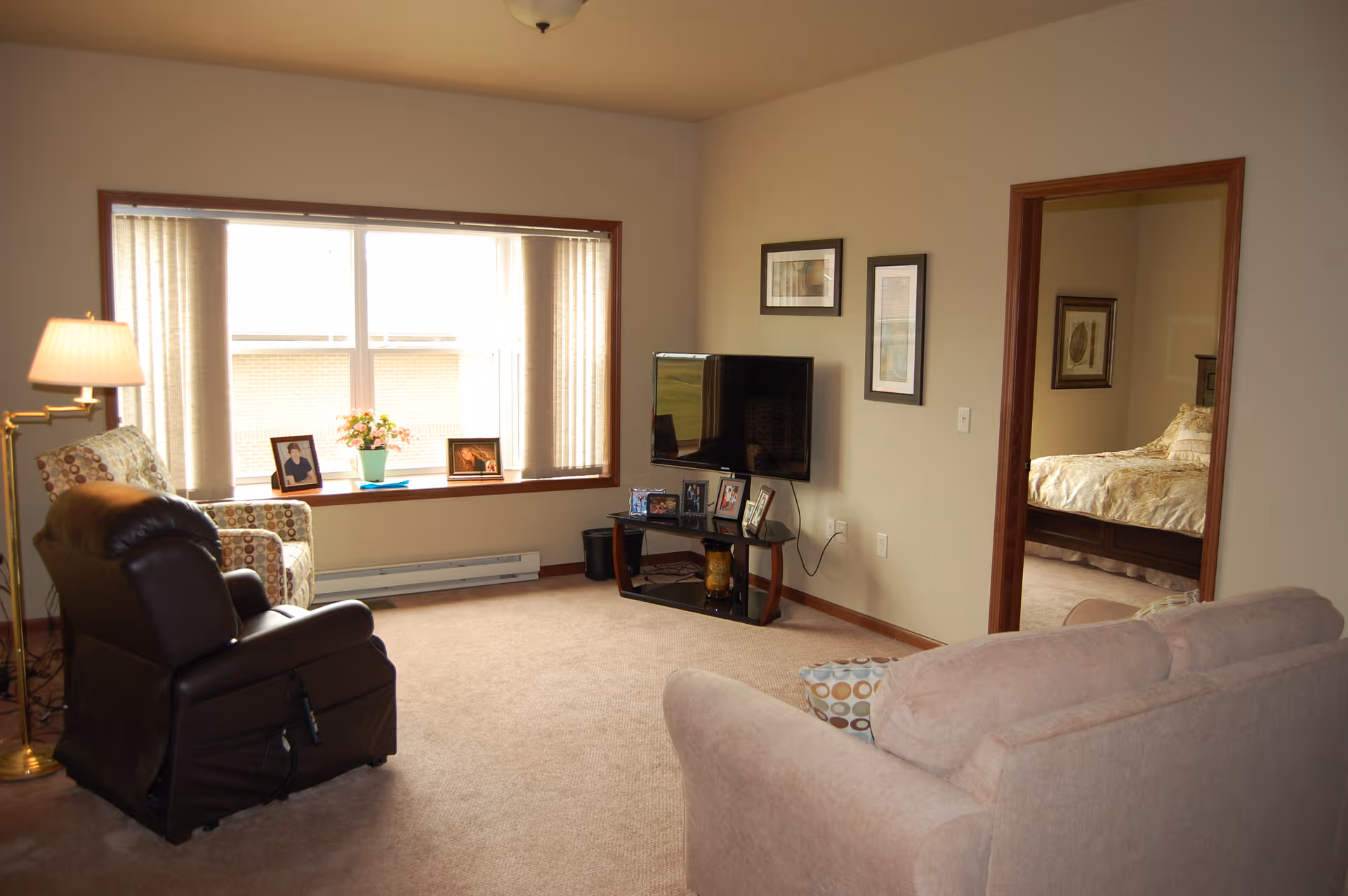 A cozy living room in an assisted living facility with a large window letting in natural light. The room features a beige sofa, a patterned armchair, a dark brown recliner, a floor lamp, and a TV mounted on a stand with framed photos. A doorway leads to a bedroom with a bed and framed artwork on the walls.