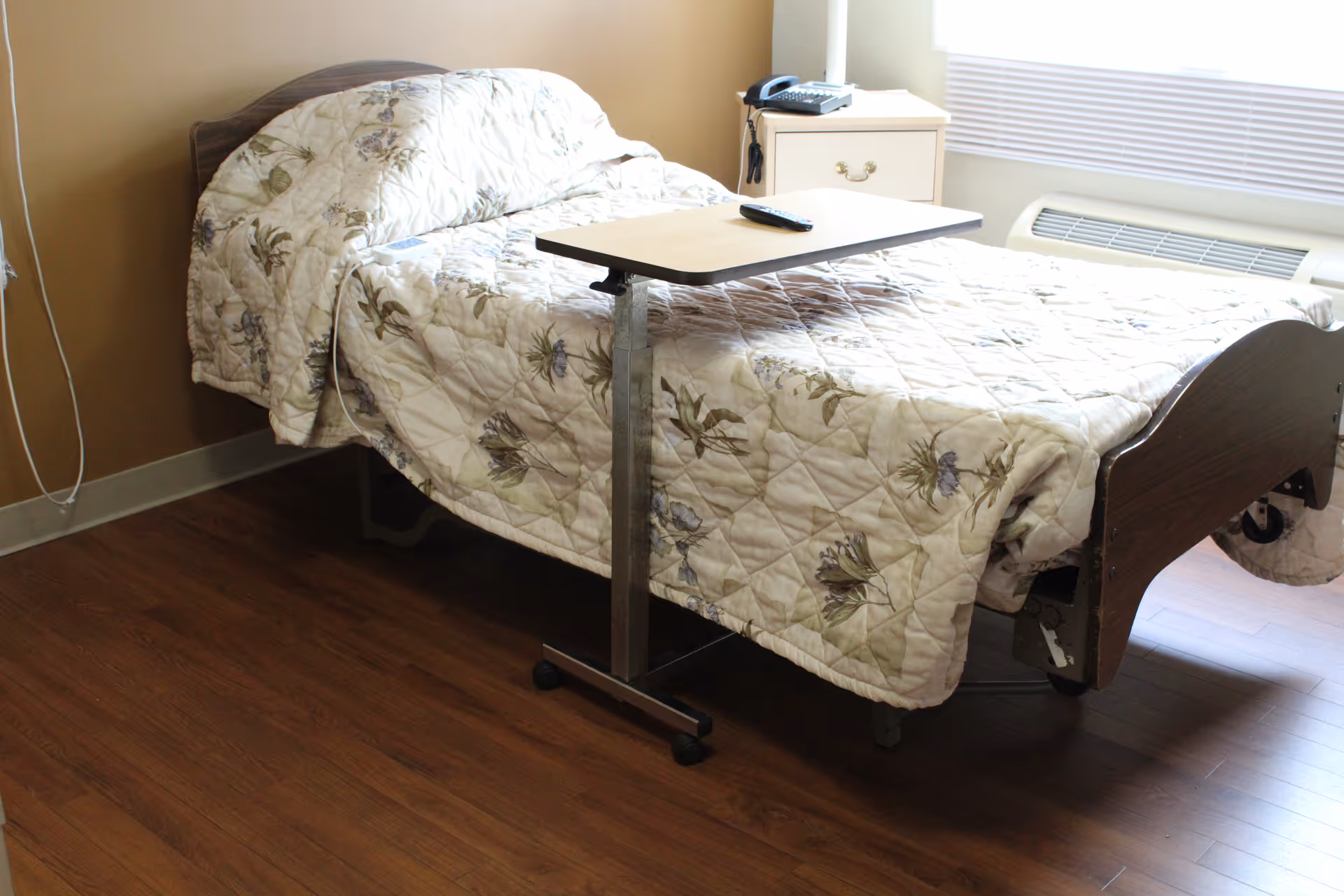 A single bed with a floral patterned quilt in a room with wooden flooring. Next to the bed is a small bedside table with a telephone on it. A rolling overbed table with a remote control on top is positioned over the bed. A window with blinds and an air conditioning unit below it is visible in the background.