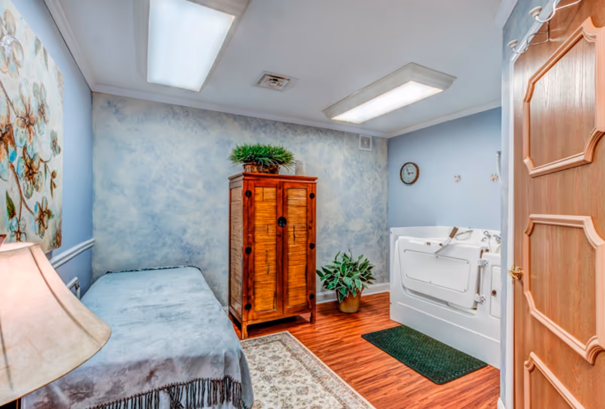 A room with a light blue wall and ceiling lights, featuring a wooden cabinet with a green plant on top, a small bed covered with a light blue blanket, a lamp on a side table, a walk-in bathtub with safety handles, a wall clock, and a potted plant on the wooden floor with a rug.