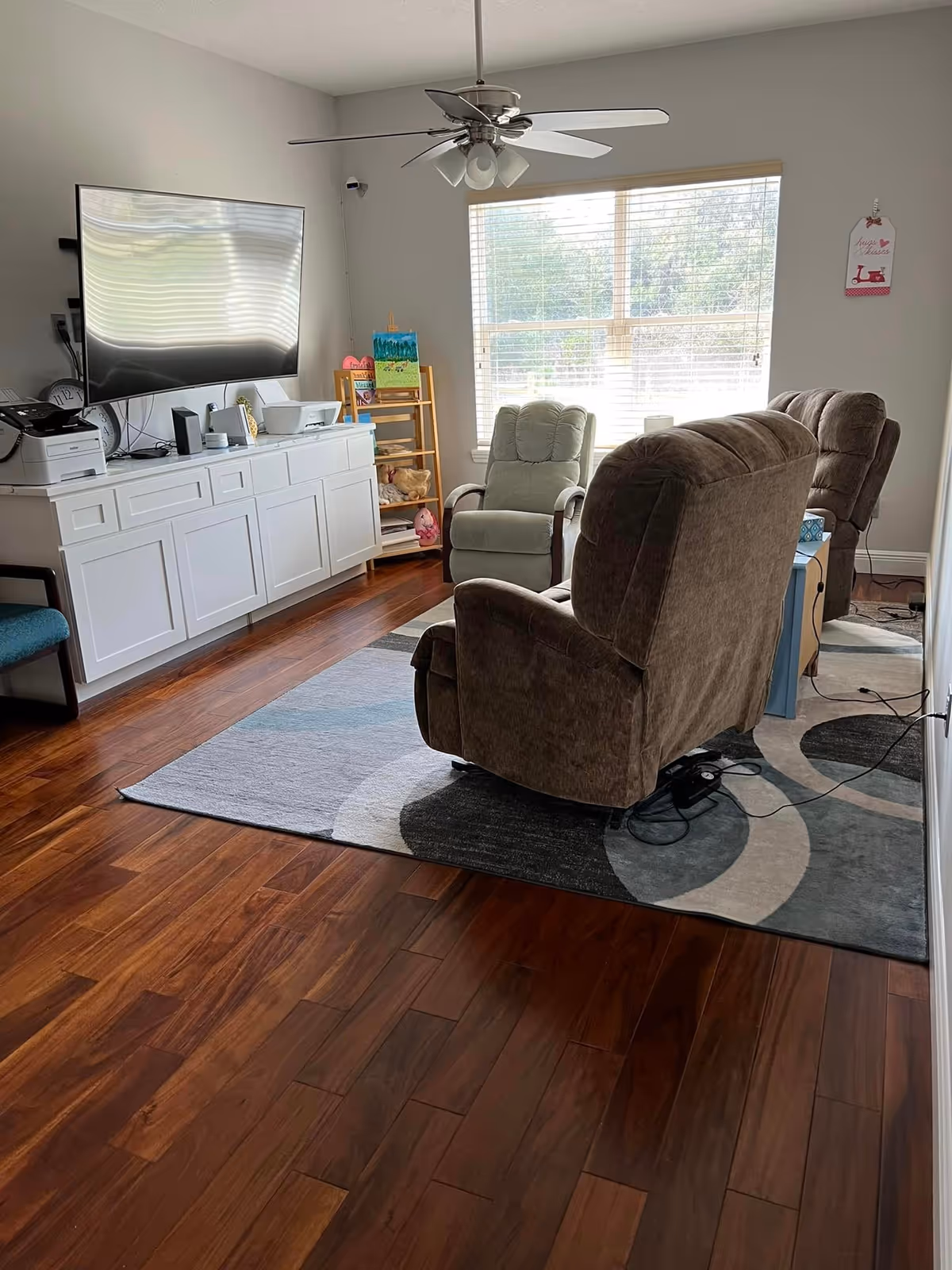 A cozy living room with wooden flooring, a large window with blinds letting in natural light, two brown recliner chairs and one gray armchair arranged on a patterned area rug. A white cabinet with a mounted flat-screen TV above it is on the left wall, along with a printer and some decorative items. A ceiling fan with lights hangs from the ceiling.