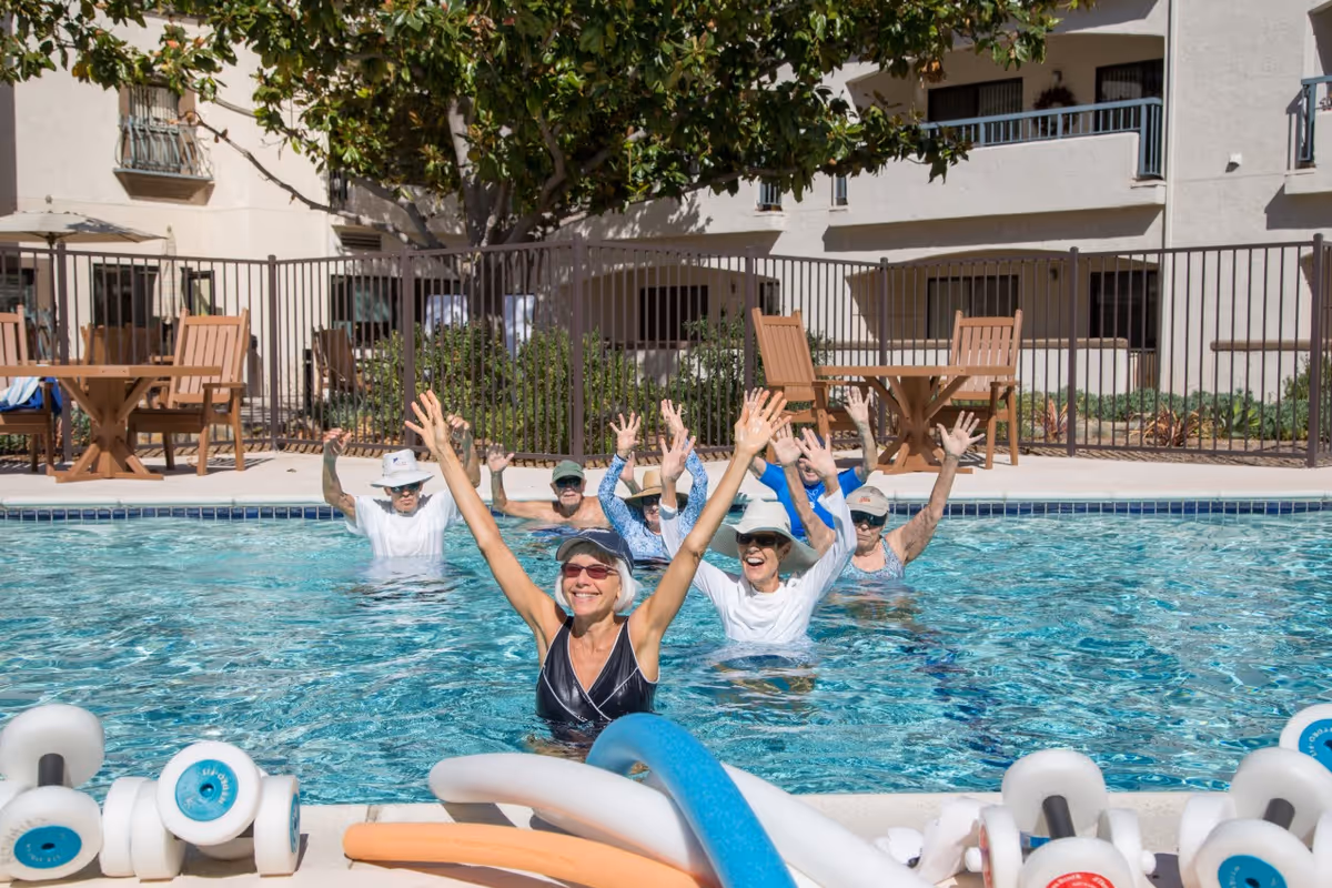 A group of elderly people participating in a water exercise class in an outdoor swimming pool, raising their arms and smiling. The pool area is surrounded by a fence, with patio tables and chairs and a building in the background.