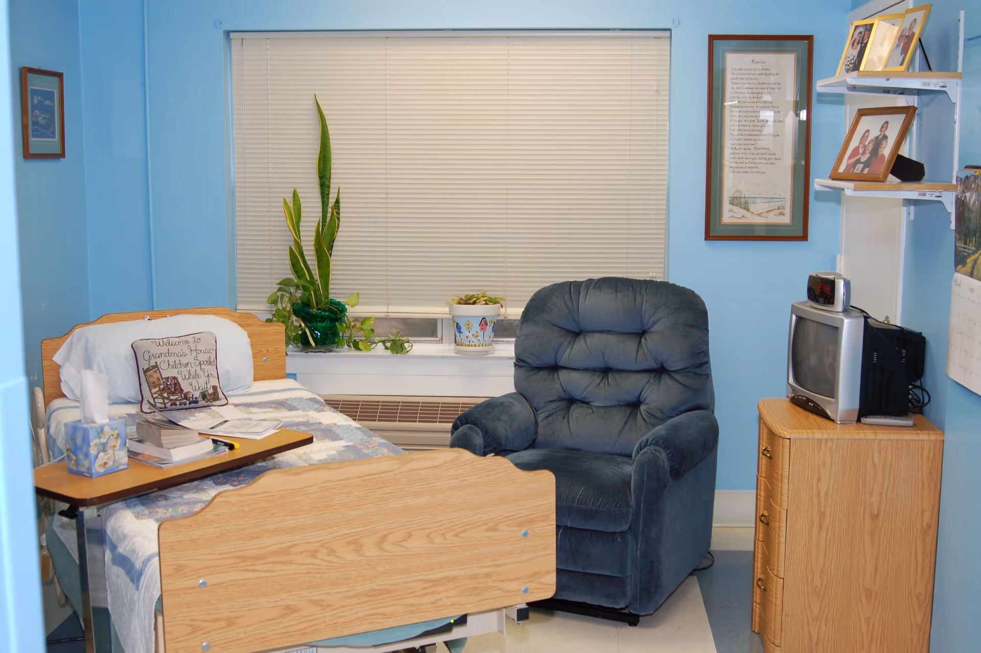 A small senior living bedroom with a hospital-style bed covered with a quilt and a pillow that reads 'Welcome to Grandma's House, Children Spoil While You Visit.' Next to the bed is a wooden tray table with books and tissues. A blue upholstered recliner chair is beside the bed. There is a window with closed blinds behind the chair and bed, with two potted plants on the windowsill. On the right side, there is a wooden dresser with a small TV and a clock on top. The walls are painted light blue and decorated with framed pictures and shelves holding family photos.