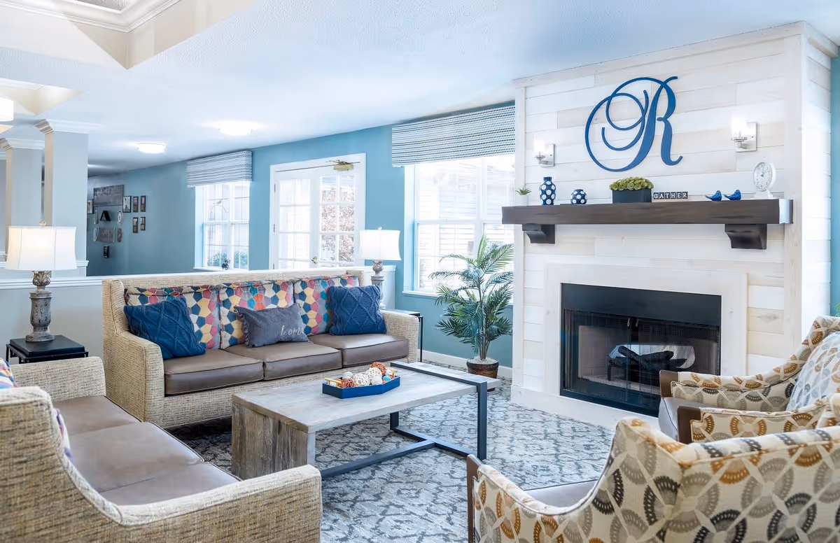 A bright and cozy living room area with a beige sofa and armchairs featuring patterned cushions. A wooden coffee table sits in the center on a patterned rug. The room has light blue walls, large windows with blinds, and a white shiplap fireplace with a dark wooden mantel decorated with vases, a clock, and a large blue letter 'R' on the wall above.