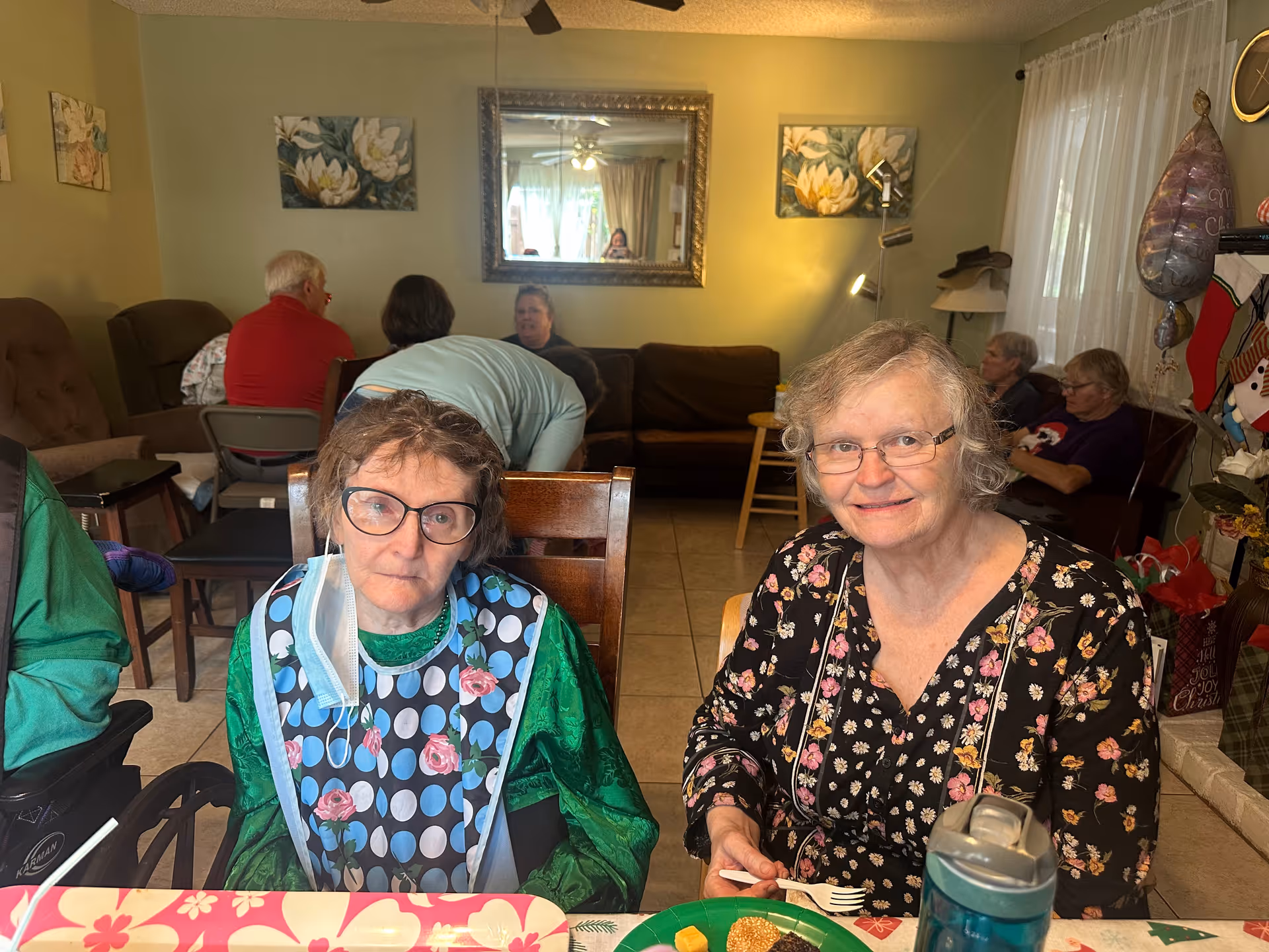 Two elderly women sitting at a table with food and drinks in a cozy living room. One woman wears glasses and a floral patterned top, smiling at the camera. The other woman wears glasses, a green shirt, and a bib with polka dots and roses, with a face mask hanging from one ear. In the background, several other elderly people are seated on chairs and couches, with floral paintings on the walls and a large mirror reflecting part of the room.