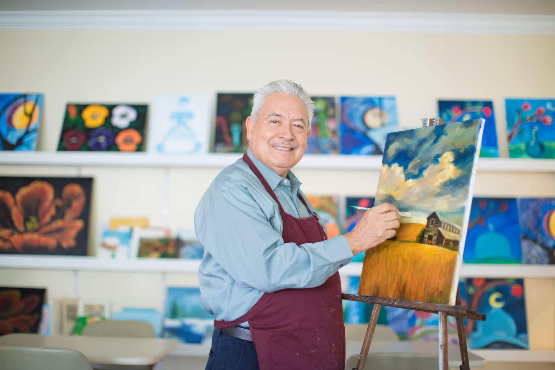 An elderly man wearing a maroon apron is painting a landscape on a canvas set on an easel. The painting depicts a field with a barn under a cloudy sky. Behind him, there are shelves filled with colorful paintings of various subjects.