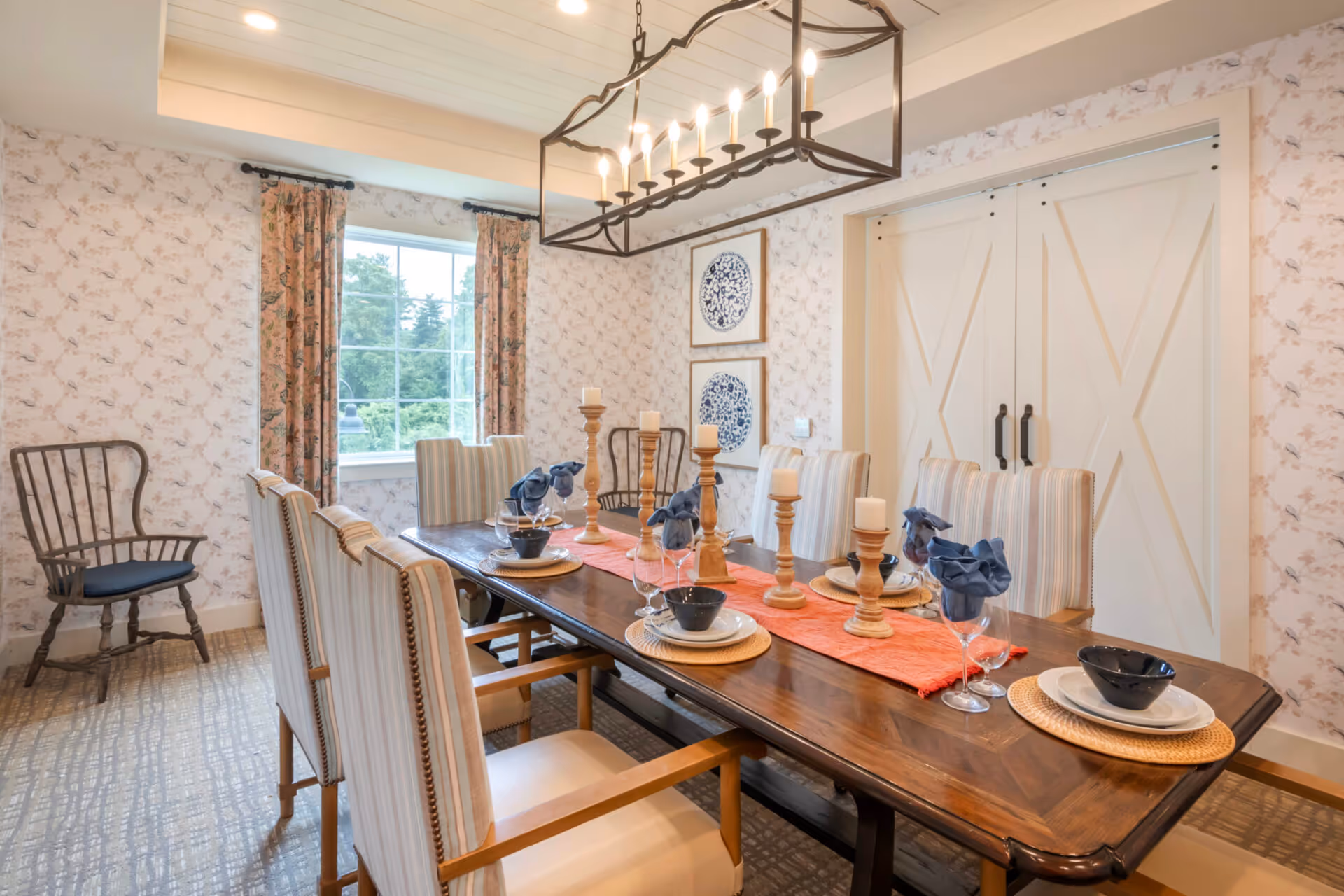 A dining room with a long wooden table set for six people. The table has a red runner, wooden candle holders with white candles, and place settings with plates, bowls, wine glasses, and blue cloth napkins. The room features floral wallpaper, a window with floral curtains, two framed blue and white decorative plates on the wall, and white double barn-style doors. There is also a wooden chair with a black cushion in the corner.