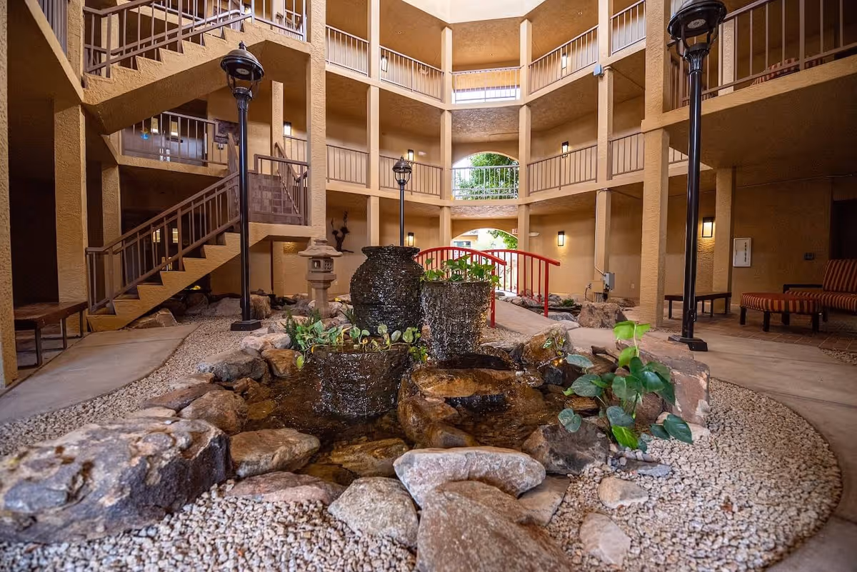 Indoor courtyard area with a small water feature consisting of three cascading pots surrounded by rocks and plants. The space is enclosed by a multi-level building with beige walls, railings, and staircases. There are two black lamp posts and seating areas with striped cushions visible on the right side.