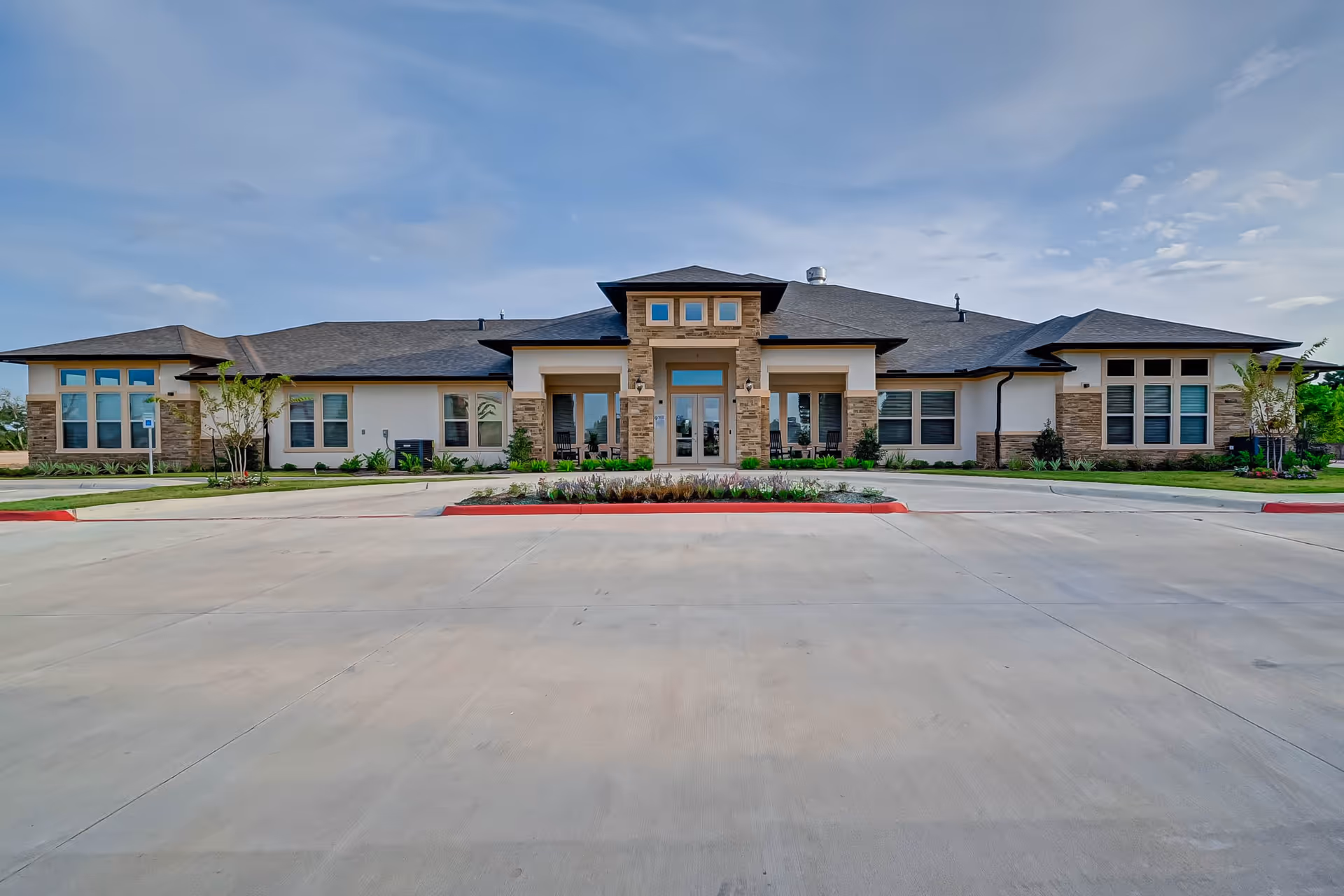 Front exterior view of a single-story assisted living and memory care facility with a wide driveway and landscaped garden beds in front. The building features large windows, a central entrance with stone accents, and a dark shingled roof under a partly cloudy sky.