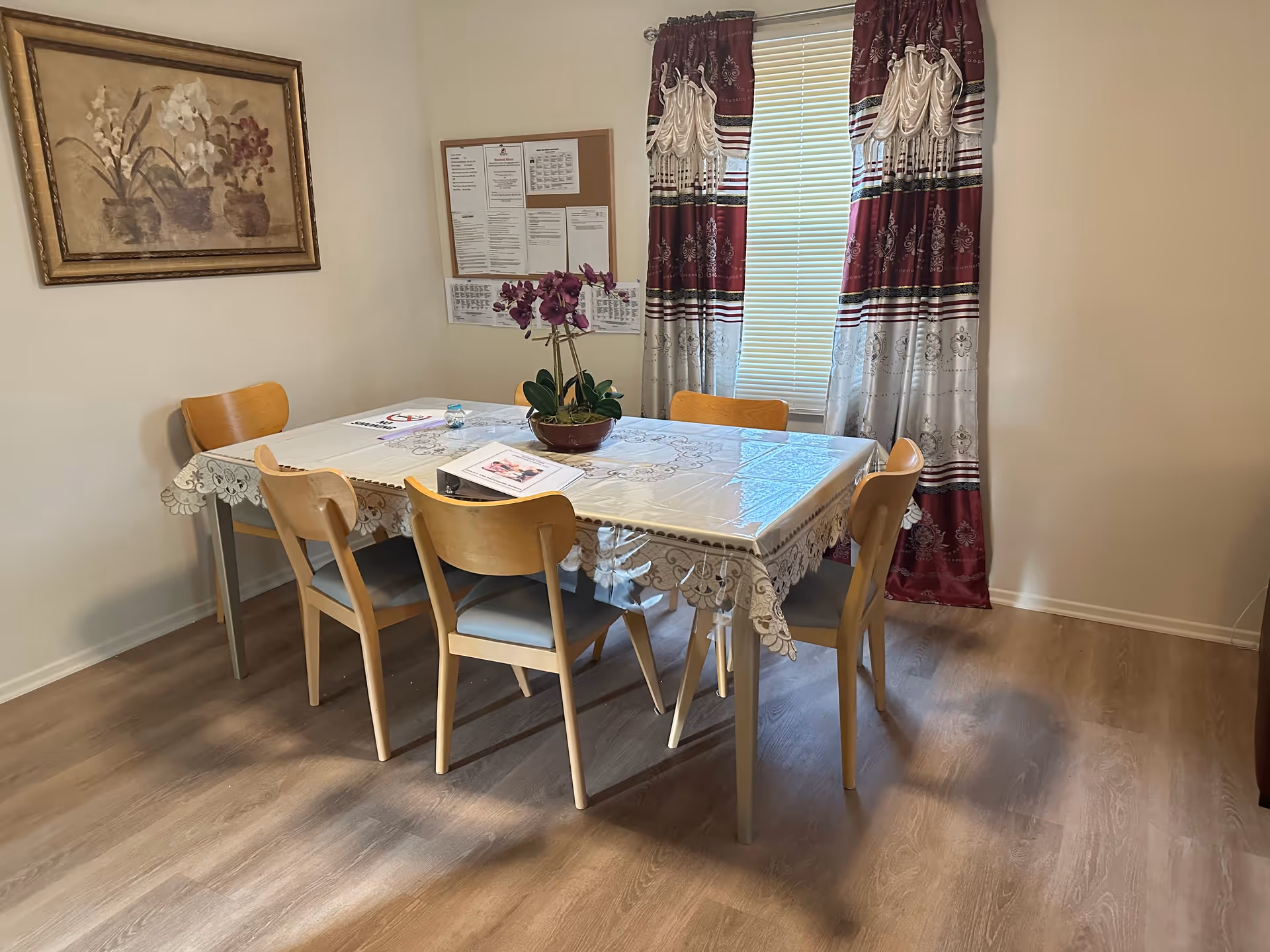 A dining room with a rectangular table covered with a lace-patterned plastic tablecloth. Six wooden chairs with light blue cushions surround the table. A potted purple orchid is placed in the center of the table. Behind the table, there is a window with red and white patterned curtains and blinds. On the wall to the left, there is a framed painting of flowers and a bulletin board with various notices.