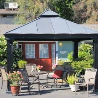 Outdoor seating area with a black metal gazebo covering a table and several chairs, surrounded by potted plants and greenery, in front of a building with red double doors and windows.