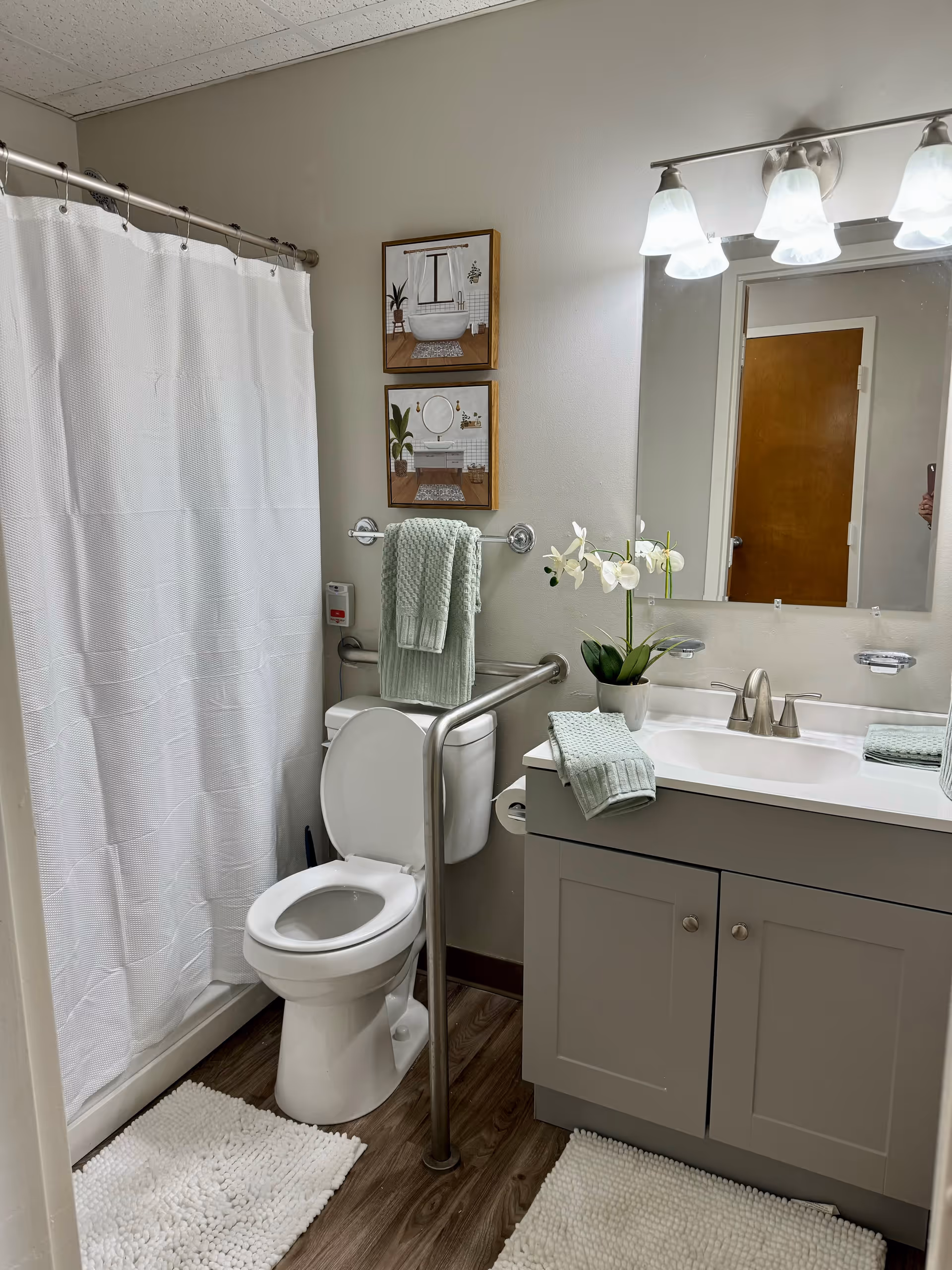 A clean and accessible bathroom with a white toilet, a white shower curtain, a gray vanity with a sink, a large mirror above the sink, and two framed pictures on the wall. There are light green towels hanging on a towel rack and placed on the vanity, a white orchid plant on the vanity, and white textured bath mats on the wooden floor. The bathroom has grab bars for accessibility.