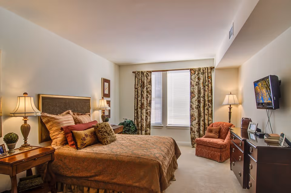 A cozy bedroom with a large bed covered in brown and patterned bedding, multiple pillows, two bedside tables with lamps, a window with floral curtains, a comfortable armchair, a dresser with framed photos and books, and a wall-mounted TV.