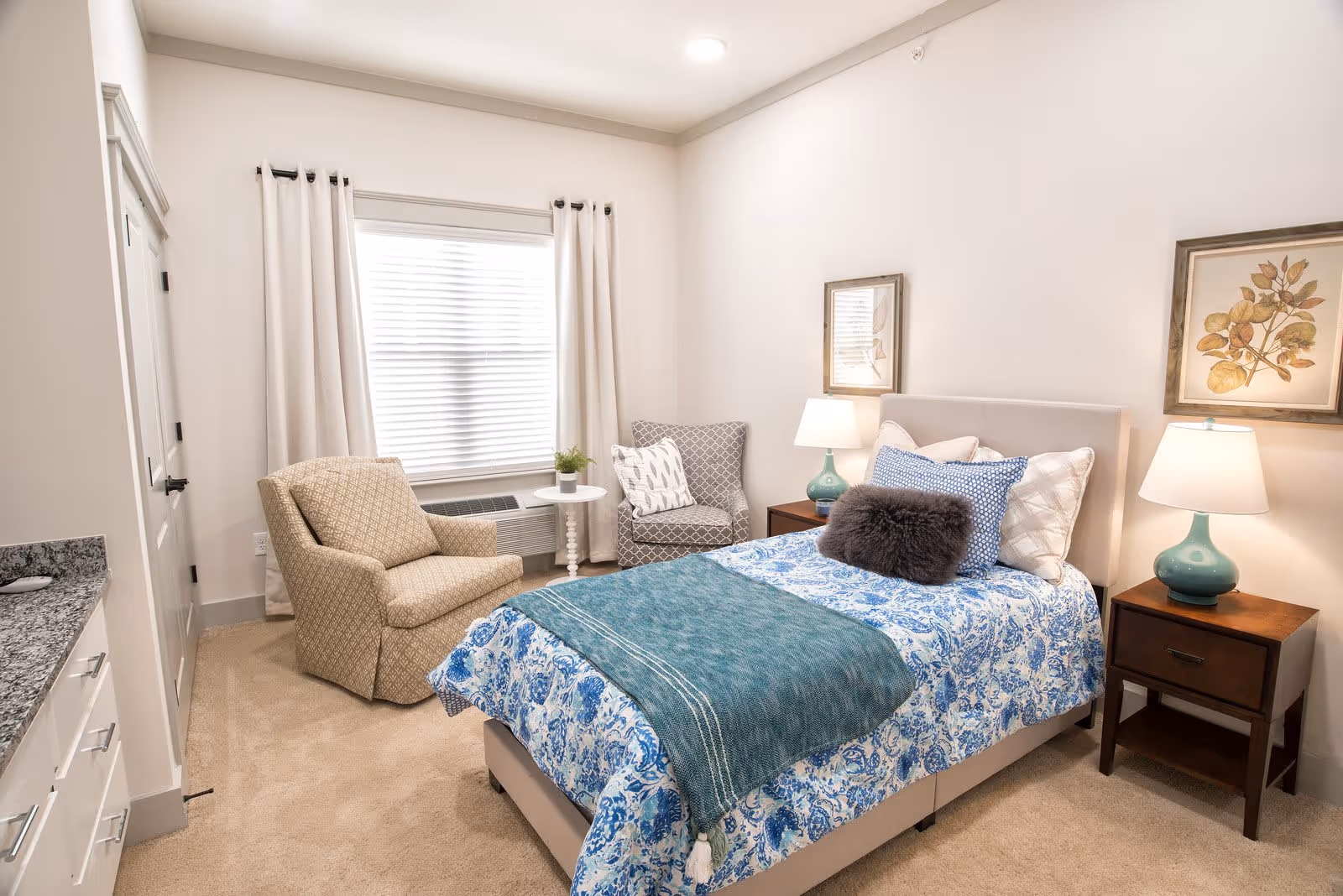 A cozy bedroom with a bed dressed in blue and white patterned bedding and a teal throw blanket. The bed has multiple pillows including a dark gray furry one. On either side of the bed are wooden nightstands with teal lamps. There are two armchairs, one beige and one gray patterned, positioned near a window with white curtains. The room has beige carpet and light-colored walls with framed botanical artwork above the nightstands.