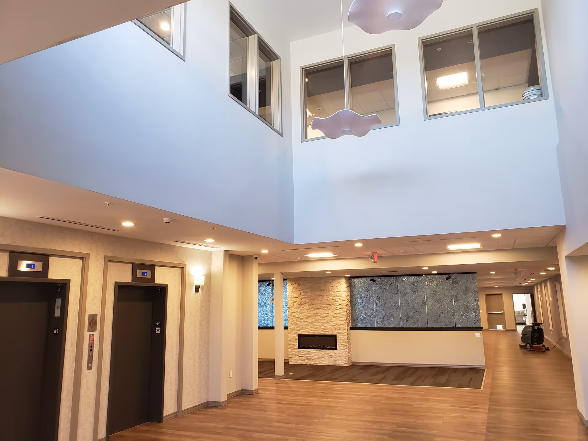 Two-story lobby atrium with elevators on the left, a fireplace feature wall, wood flooring, and upper-level windows.