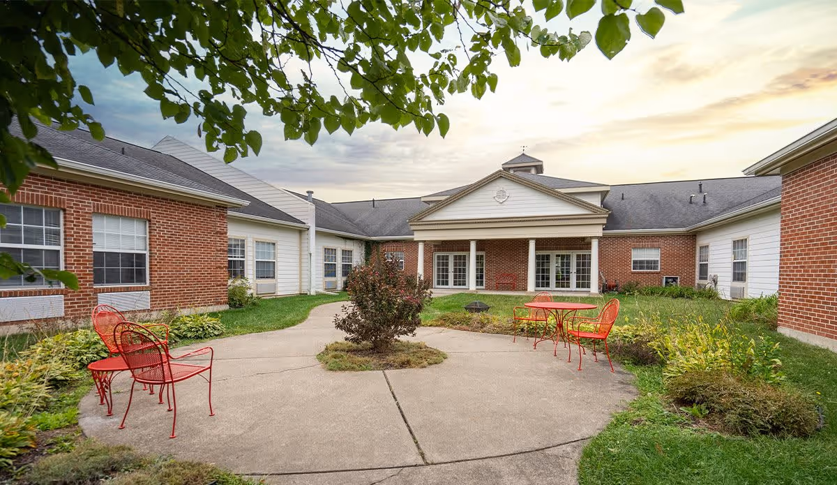 Outdoor courtyard area of Walnut Ridge Assisted Living and Memory Care with a concrete pathway, red metal chairs and tables, surrounded by green grass, bushes, and a brick building with white trim under a partly cloudy sky.