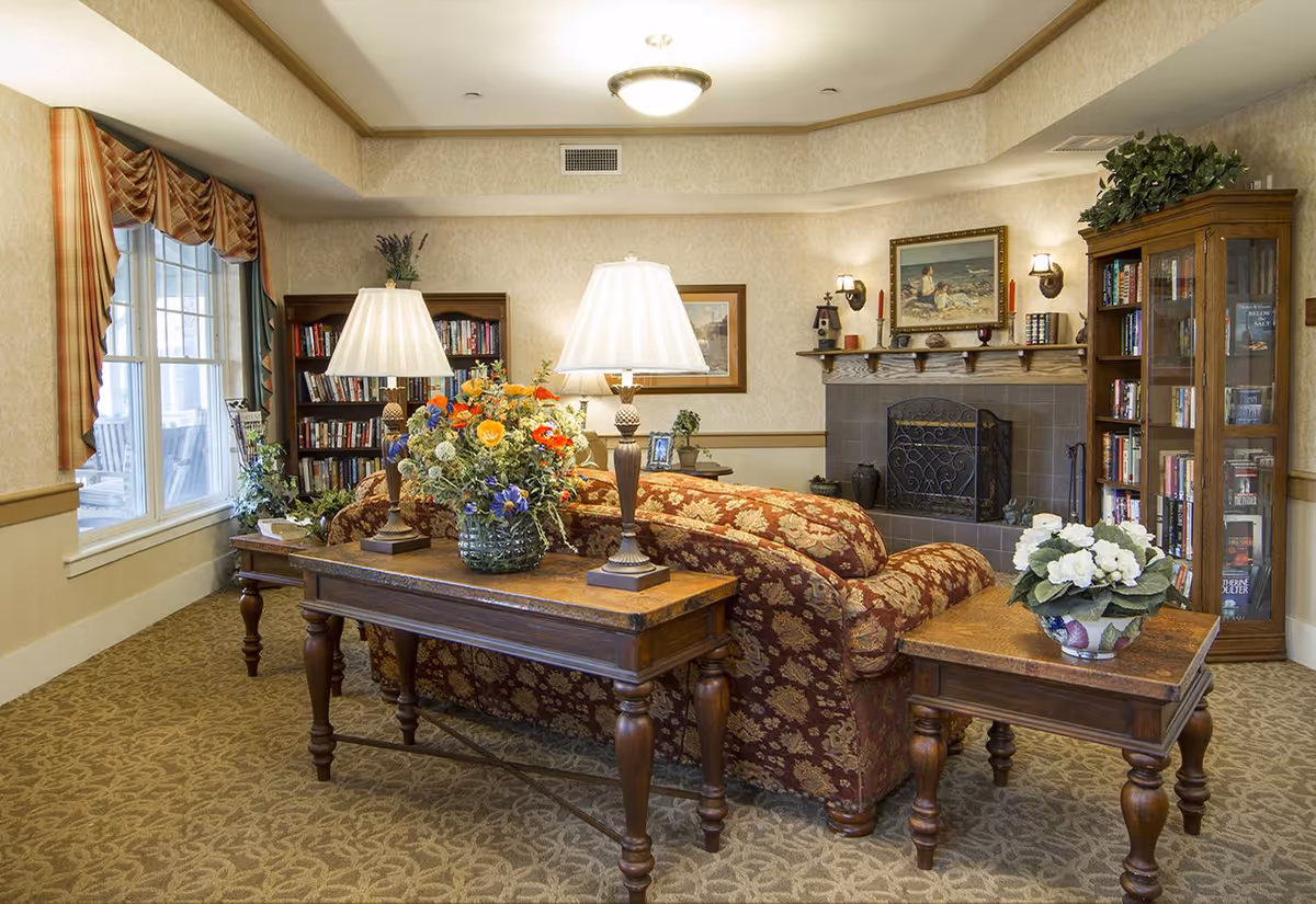 Cozy communal living room with a patterned sofa, wooden tables topped with lamps and floral arrangements, bookshelves and a fireplace.